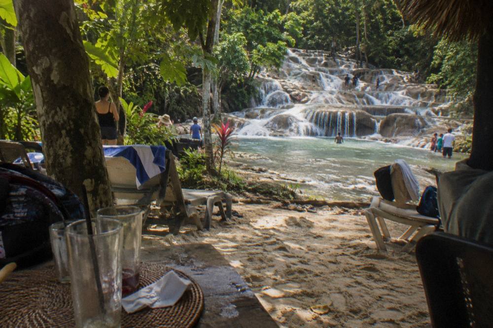 Golden light at a Jamaican beach club, woven placemats and tall glasses on pale sand, terraced falls behind