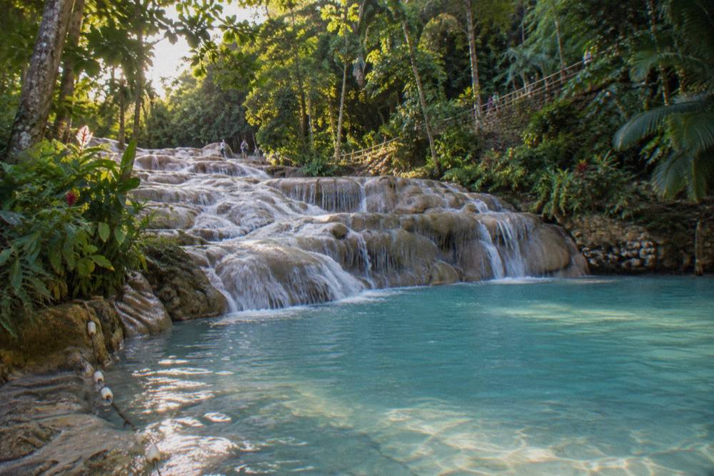 Turquoise natural pool at the base of a terraced waterfall with swimmers standing in shallow water on smooth rock