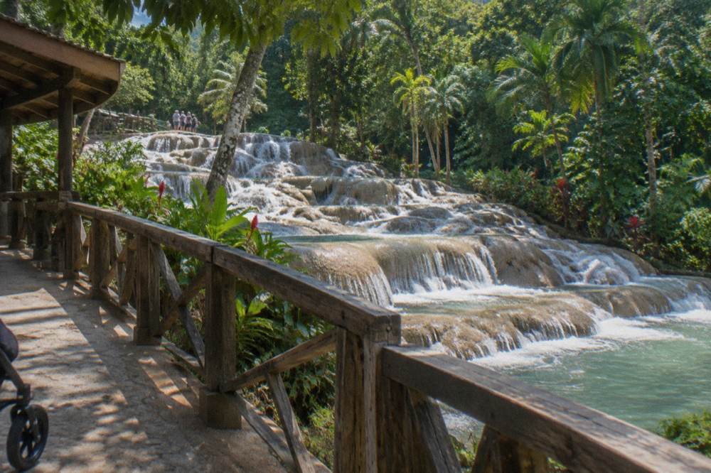A paved viewing path and wooden deck running beside a terraced waterfall with tropical trees overhead