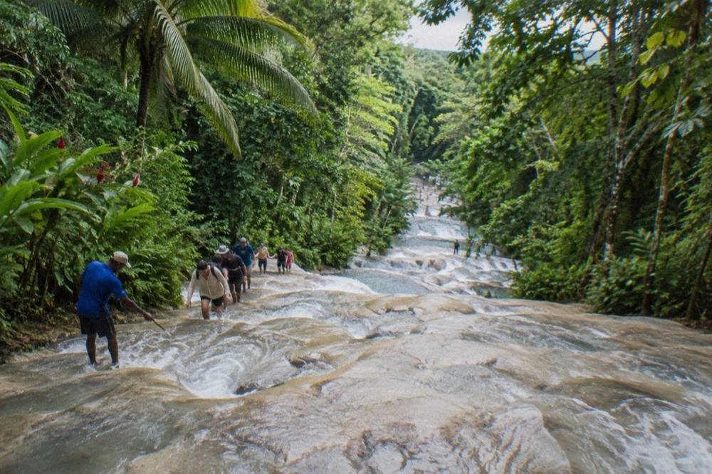 Visitors forming a chain in the water on wide limestone steps of a waterfall surrounded by forest