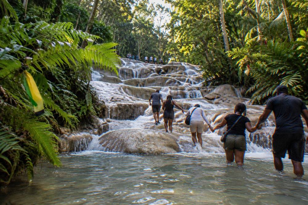 Visitors forming a chain in the shallow pools of a terraced waterfall, forest on both banks