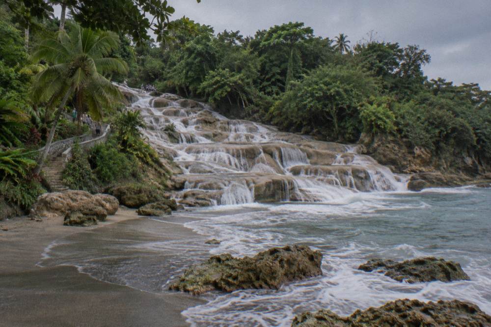 Sandy beach at the base of a waterfall where fresh water meets the Caribbean sea under an overcast sky