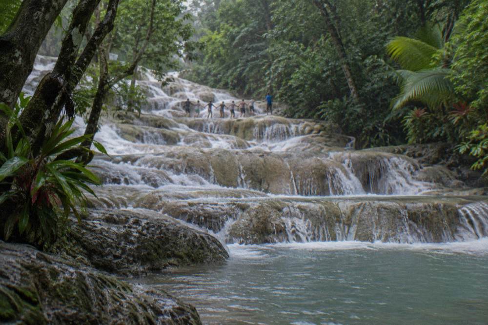 Visitors in swimwear forming a chain as they climb a stepped waterfall beside a guide