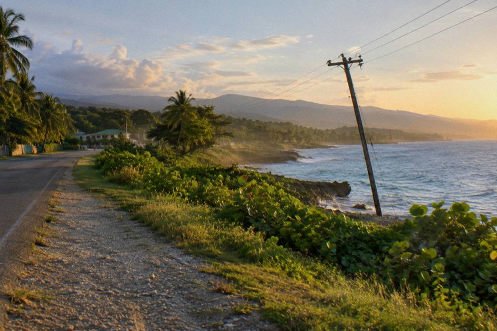 Golden light raking a green shoreline toward distant blue hills, dust hanging by a coastal road, leaning pole