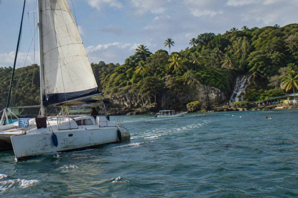 Sailing catamaran with full sails along golden limestone cliffs and palms, wake fanning across calm blue water