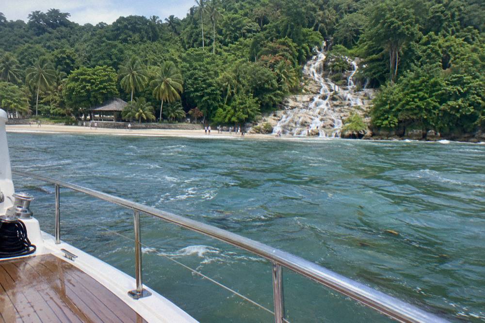 From a moored catamaran deck across calm turquoise water toward a sandy beach and terraced limestone falls