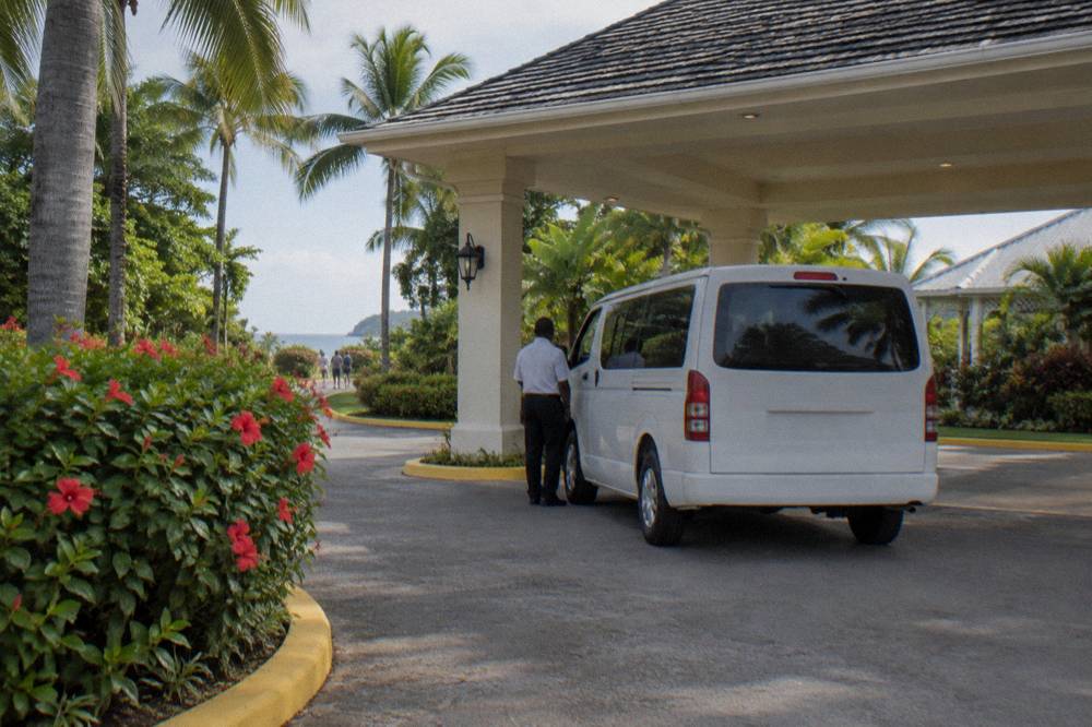 Plain white minivan under a covered entrance with hibiscus and palms, driver standing beside with back turned.