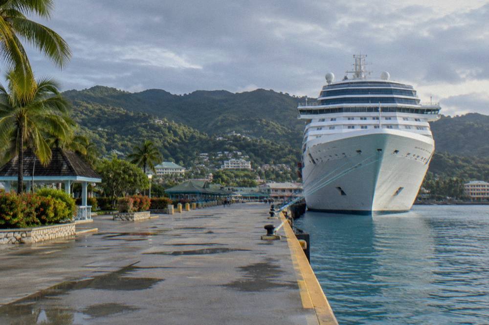 White cruise ship docked at a Caribbean port town with green hills behind