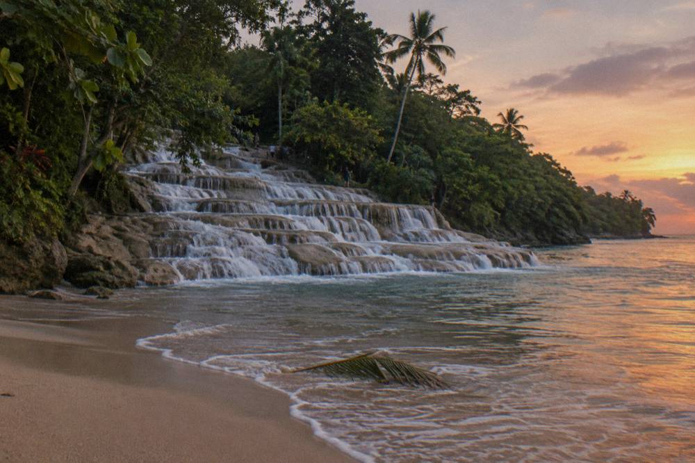 Dusk at a beach where terraced falls spill onto golden sand, foam on the wet shore, a palm frond in shallows