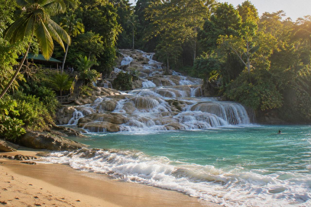 Turquoise terraces of a wide waterfall cascading toward a sandy beach under a bright sky