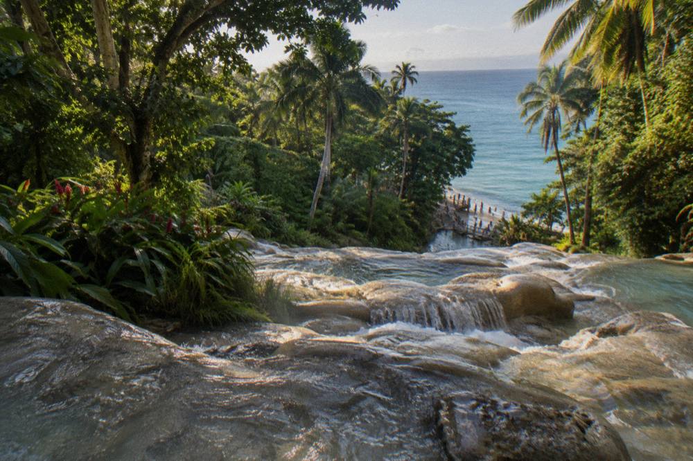 Waterfall terraces stepping down through dense green forest toward a sandy beach