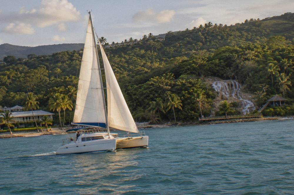 Catamaran with raised sails cutting across calm water along the Jamaican coast
