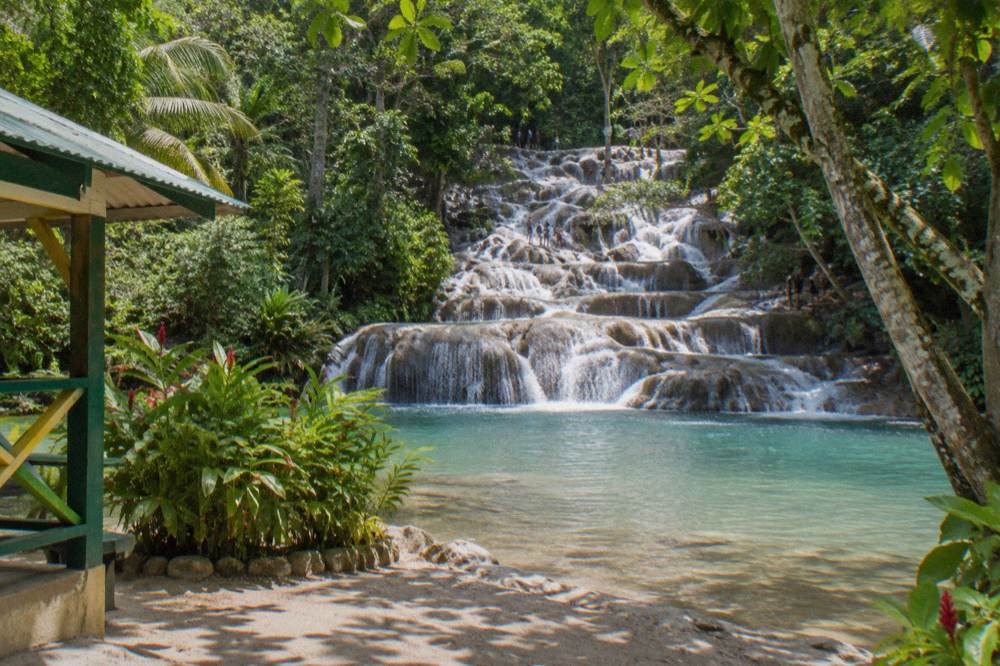 Sandy beach at the base of a waterfall with sun hats and towels on a bench