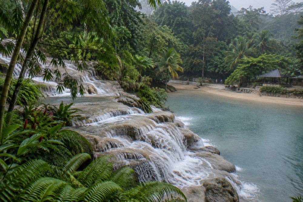 Waterfall terraces stepping down through tropical foliage toward a calm sandy beach